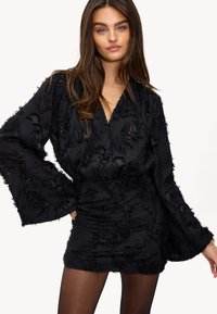 Zwarte jurk van getextureerde stof, met een diepe V-hals, lange klokmouwen en gerimpelde detailing. Accenten van rafelige randen overal.