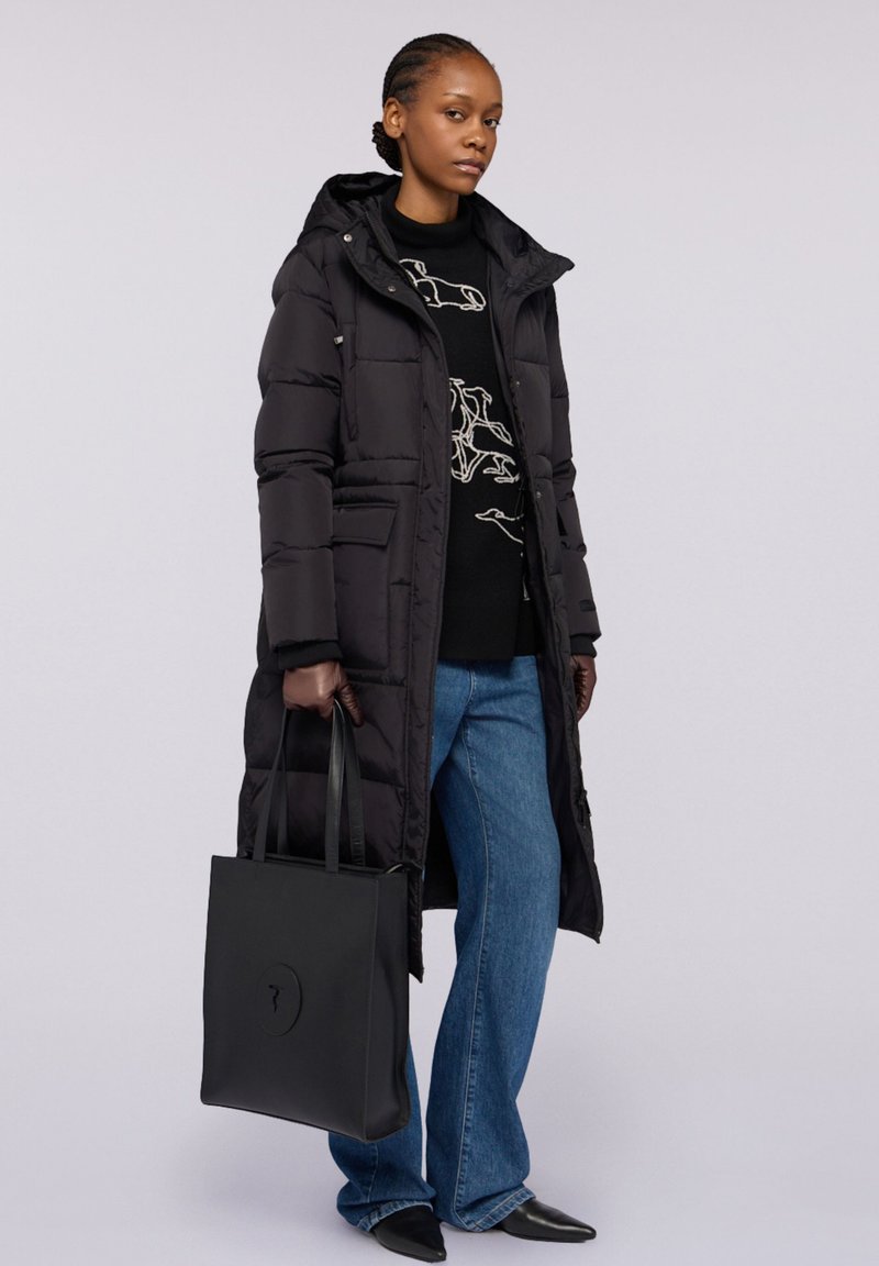 Svart vadderad lång kappa med huva, bärs över en svart tröja med ljus design, kombinerad med blå utsvängda jeans och en svart tygväska.