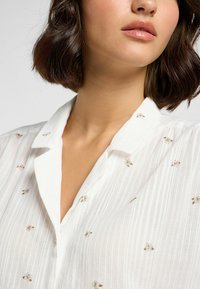 Camisa blanca ligera con finas rayas verticales y pequeño bordado floral en rosa y verde. Cuello clásico y cierre frontal con botones.