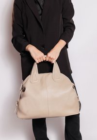 Persona in giacca nera che tiene una grande borsa di pelle beige con due manici su uno sfondo chiaro e semplice.