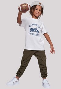 White graphic t-shirt with blue motorcycle print, olive green cargo pants, and white sneakers with gray accents. Holding a brown football.