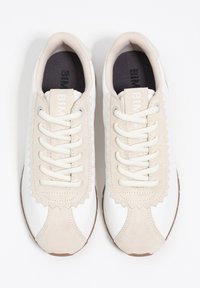 Białe i beżowe sneakersy z teksturowaną cholewką, falbankami na języku i kremowymi sznurowadłami. Brązowa podeszwa.