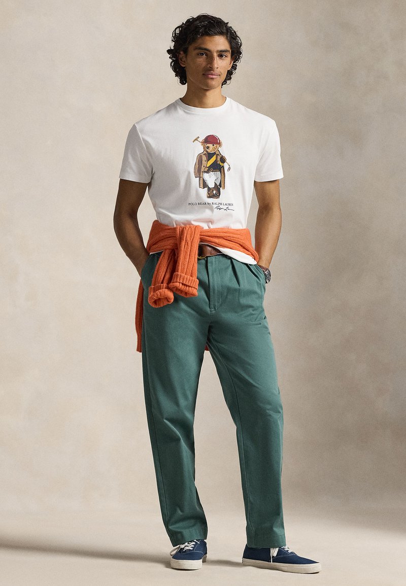 Weißes Baumwoll-T-Shirt mit einem Grafikdruck, orangefarbener Strickpullover um die Taille gebunden, grüne Hose und marineblaue Schuhe. Lässiges Outfit.