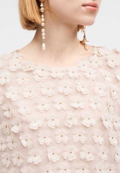 Απαλό ροζ μπλούζα με στρώσεις από λουλουδένιες εφαρμογές και μεταλλικές χάντρες, συνοδευόμενη από κρεμαστά σκουλαρίκια με μαργαριτάρια και χρυσά στοιχεία.