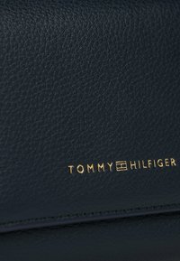 Close-up of textured dark leather with gold Tommy Hilfiger logo embossed on the surface near stitched edge.