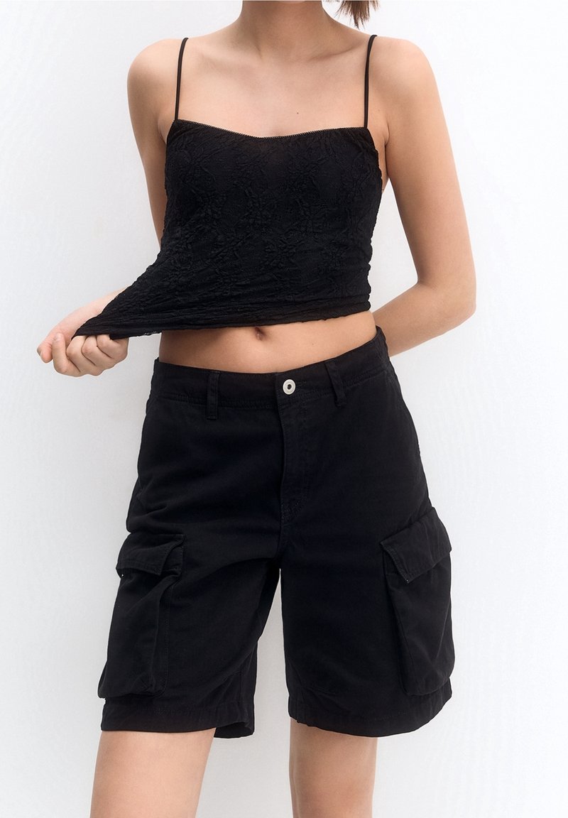 Camiseta de encaje negra con tirantes finos, combinada con pantalones cortos de carga negros que cuentan con bolsillos laterales y un ajuste relajado.