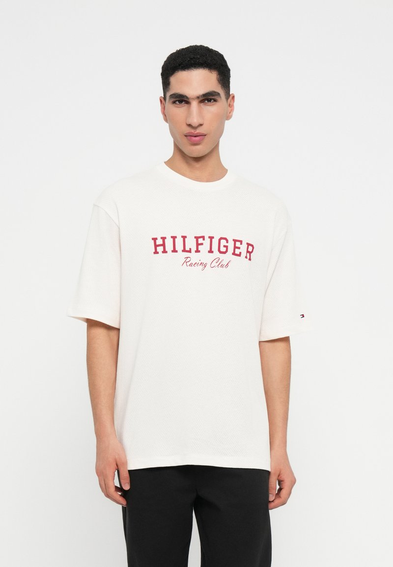 Νεαρός άνδρας φοράει ένα λευκό κοντομάνικο μπλουζάκι με την ένδειξη "HILFIGER Racing Club" σε κόκκινες γράμματα, σε συνδυασμό με μαύρο παντελόνι, μπροστά από λευκό φόντο.