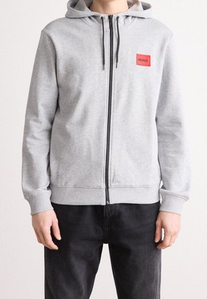 Person trägt eine hellgraue Kapuzenjacke mit Reißverschluss, Kapuze und schwarzen Kordeln, mit einem roten "HUGO"-Aufnäher auf der Brust.