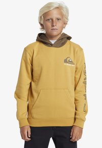 Gul huvtröja med en brunfärgad innerventil, framficka och tryckt logotyp på bröstet och ärmen. Mjuk tygkvalitet och avslappnad passform.