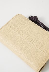 Portefeuille en cuir beige avec texture granuleuse, nom de marque embossé "Coccinelle" et détail de tirette en cuir tressé violet.