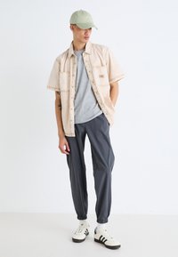 Chemise beige clair à manches courtes sur un t-shirt gris, associée à un pantalon fuselé gris foncé et des baskets blanches avec des rayures noires. Casquette verte incluse.