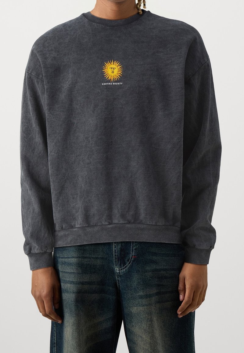 En person som bär en mörkgrå sweatshirt med en gul solsymbol och texten "Kaotiko Society" på bröstet, tillsammans med blekta blå jeans.