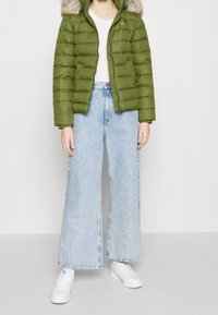 Giacca puffa verde oliva con cappuccio foderato in pelliccia, abbinata a jeans azzurri a gamba larga e sneakers bianche. Texture liscia, chiusura con zip.
