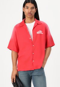 Camisa roja de manga corta con un diseño de botón y logo bordado en el pecho. Lleva una camiseta blanca debajo y está combinada con jeans azul.