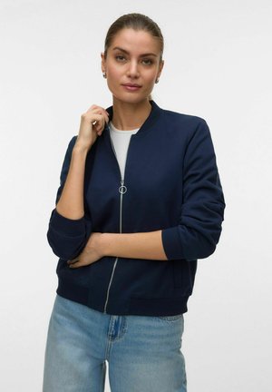 Donna con capelli tirati indietro che indossa una giacca blu navy con zip sopra una maglietta bianca e jeans azzurri, in piedi davanti a uno sfondo semplice.