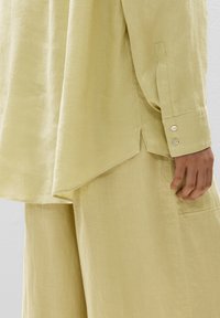 Camicia di lino e pantaloni a gamba larga in giallo chiaro. Presenta un orlo curvo, maniche lunghe con bottoni e un tessuto leggero e testurizzato.