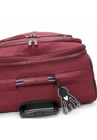 Maroon suitcase with dual zippered compartments, a retractable handle, rubber wheels, and a black monkey-shaped tag. Smooth fabric texture.