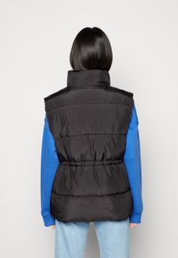 Czarna ocieplana kamizelka z wysokim kołnierzem i elastycznym pasem, o gładkiej fakturze i pikowanym wzorze. Noszona na niebieską koszulę z długim rękawem.