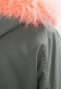 Grüne Jacke mit einem flauschigen, rosafarbenen Kunstpelzkragen. Der Stoff ist glatt mit einer matten Oberfläche, die ein lässiges und modernes Design unterstreicht.