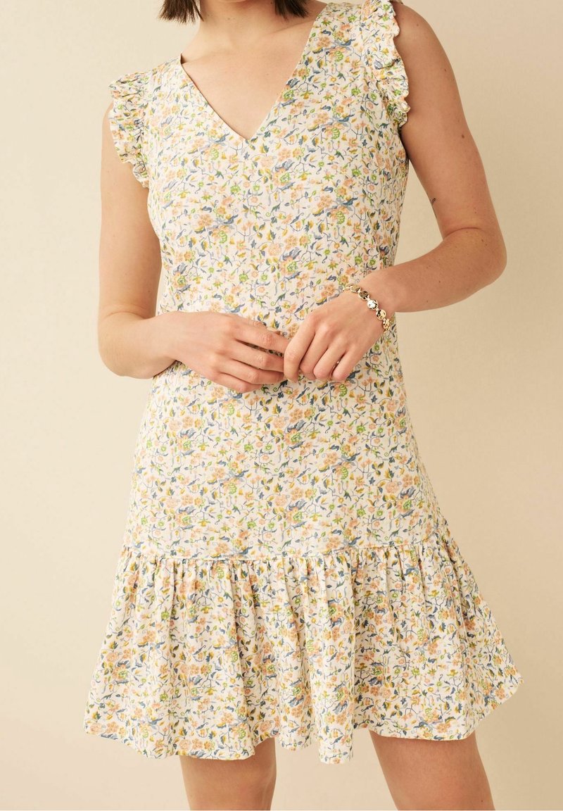 Floral jurk met een V-hals, gerimpelde mouwen en een uitlopende zoom. Kenmerkt zich door een witte basis met gele, blauwe en groene bloemenpatronen. Zachte stof.