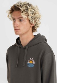 Grijze hoodie met een voorzak, met een kleurrijk cirkelontwerp van een zon en golven. De stof lijkt zacht en textuur te hebben.