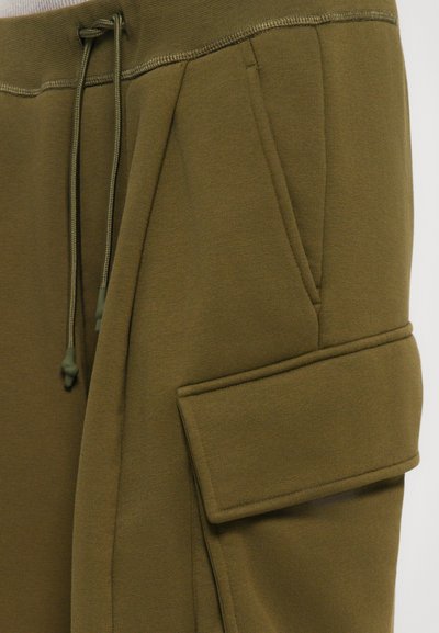 Olive green sweatpants with side drawstring, angled pockets, and large flap pocket on one thigh, shown in close-up view.