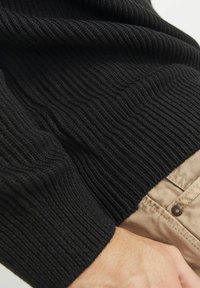 Schwarzer gerippter Pullover aus gestricktem Stoff mit strukturierten Details, eng anliegenden Ärmeln und einem Saum, der über hellfarbige Hosen mit einem Knopf überlappt.