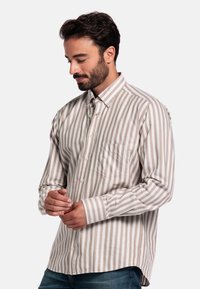 Man die de manchet van een beige en witte verticaal gestreepte button-up shirt aanpast, met op de achtergrond een effen witte achtergrond, gekleed in een blauwe jeans.