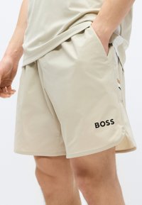 Svetlo bež kratke hlače iz lahkotnega materiala z bočnimi žepi. Imajo črno logotip "BOSS" in subtilne oblikovalske poudarke na straneh.