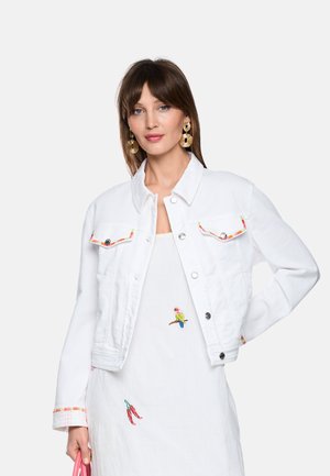 Frau trägt eine weiße, kurz geschnittene Jacke mit buntem Besatz und ein weißes Kleid mit Papagei- und Chilischoten-Stickerei, ergänzt durch große goldene Ohrringe.