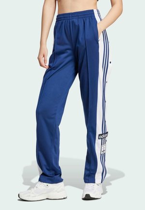 Vrouw die blauwe en witte Adidas trainingsbroek met zijstrepen en witte sneakers draagt, staand met één hand in de zak.