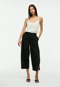 Donna che indossa un top in pizzo bianco, pantaloni neri a gamba larga e cropped con spacchi laterali, e sandali neri con punta aperta, in piedi con una mano in tasca.