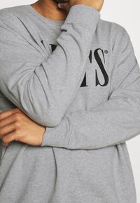 Sudadera de manga larga gris con cuello redondo, presenta texto impreso en negro en el frente. La tela parece suave y tiene un ajuste casual.