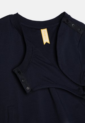 Haut en coton bleu marine avec un col rond, fermeture à bouton-pression à l'épaule et une étiquette en tissu jaune portant la mention "FRI BOO". Texture douce.