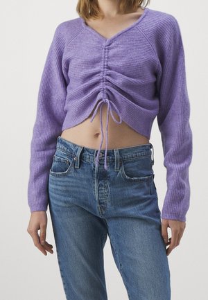 Woman wearing a cropped, ruched purple knit sweater with long sleeves and high-waisted blue jeans against a plain background.