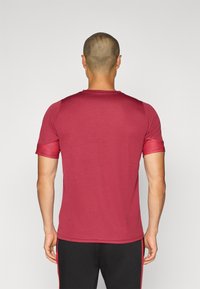 Bordeaux színű sportpóló rövid ujjakkal, kerek nyakkal és texturált válldíszítéssel. Az anyag könnyűnek és simának tűnik.