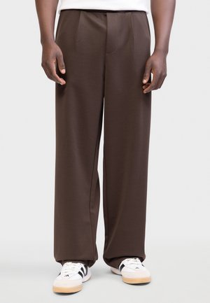 Vyras dėvi laisvus rudus kelnes ir baltus sportinius batelius su juodomis juostelėmis, stovintis priešais šviesų, vienspalvį foną.