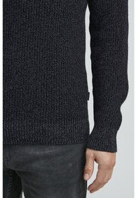 Dunkel gestrickter Pullover mit einem strukturierten, gerippten Muster und schmalen Bündchen. Mit einem Seitenschild ausgestattet, kombiniert mit dunkelgrauen Hosen.