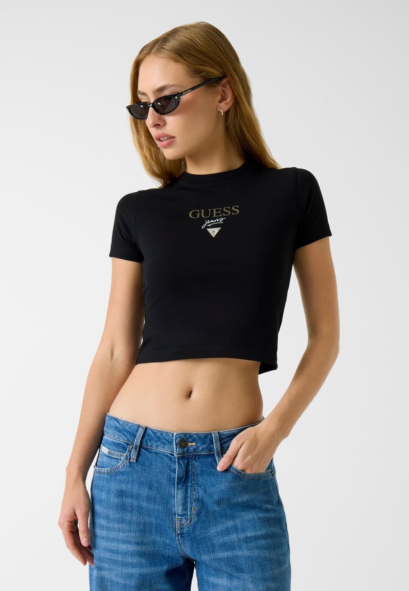 Czarny krótki t-shirt wykonany z jerseyu z złotym logo "GUESS", zestawiony z niebieskimi dżinsami. Krótkie rękawy, dopasowany fason.