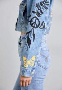 Džinsinis švarkas su juodais grafiškais lapų, taikos simbolio ir geltonų drugelių dizainais ant rankovių. Prigludęs fasonas, šviesiai mėlyna spalva.