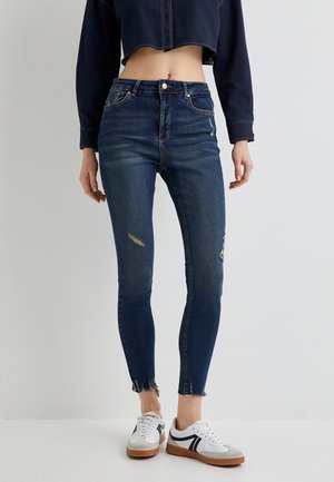 Jean taille haute bleu foncé avec une coupe slim, genoux déchirés, ourlet effiloché et design à cinq poches, associés à des baskets blanches.