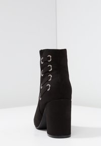 Botines de ante negro hasta el tobillo con un alto tacón cuadrado, que cuentan con ojetes plateados y detalle de cordones en la parte trasera. Textura suave; diseño elegante.