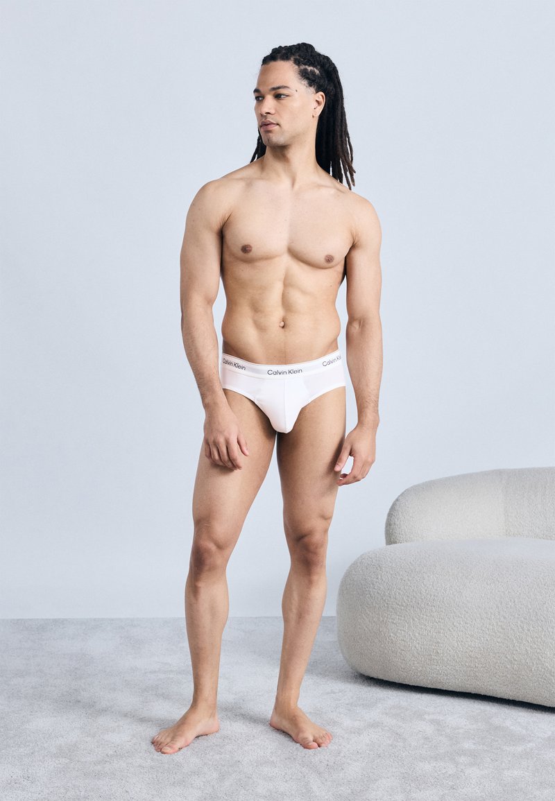 Μυώδης άντρας με κοτσίδες ντυμένος με λευκά εσώρουχα Calvin Klein στέκεται ξυπόλυτος σε παρκέ δίπλα σε καμπυλωτό, υφασμένο λευκό καναπέ.