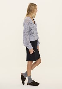 Helle lavendel gestrickte Strickjacke mit einem offenen Spitzenmuster, Knopfverschluss und gerippten Bündchen, kombiniert mit einem schwarzen, figurbetonten Kleid und grauen Socken.