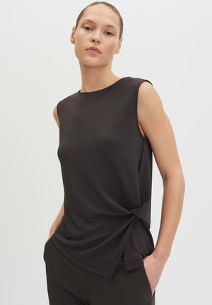 Femme aux cheveux attachés portant un haut noir sans manches avec un détail de nœud sur le côté et un pantalon noir assorti, mains dans les poches, fond neutre.