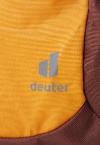 Tecido amarelo e castanho com um acabamento texturizado, apresentando um logótipo "deuter" em cinza. O design mostra uma transição suave entre as cores.