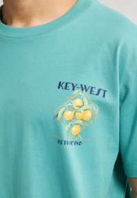Turkoosi puuvillapaita, jossa on graafinen kuvasto keltaisista orangesista vihreillä lehdillä sekä tekstit "KEY-WEST" ja "PETROLIND" tummansinisellä.