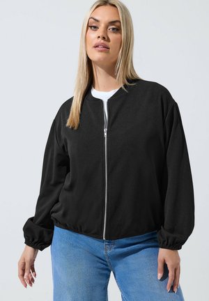 Woman with straight blonde hair wearing a black zip-up jacket over a white shirt and blue jeans, standing against a light background.