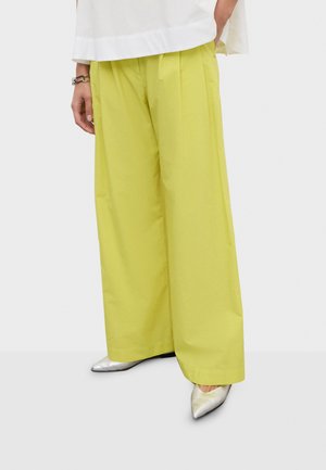 Osoba ubrana w limonkowe spodnie szerokie na nogawki, srebrne spiczaste buty i biały top, stojąca na tle jednolitego jasnego tła.