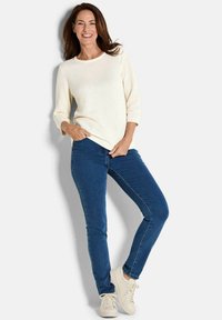 Heller, strukturierten Pullover mit drei Viertelärmeln, kombiniert mit dunkelblauen Skinny-Jeans und weißen Sneakers. Model posiert mit den Händen in den Hüften.